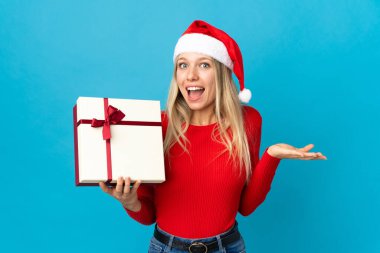 Noel şapkalı kadın elinde hediyesiyle mavi arka planda şok olmuş bir yüz ifadesiyle izole edilmiş.