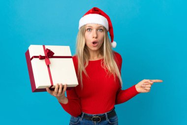 Noel şapkalı kadın mavi arka planda bir hediye tutuyor. Şaşırmış ve işaret ediyor.