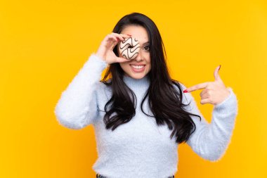 Genç Kolombiyalı kız izole edilmiş sarı arka planda elinde donut ve mutlu