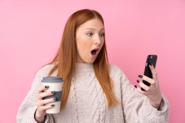Genç kızıl saçlı, soyutlanmış pembe arka planda, elinde kahve ve cep telefonu olan bir kadın.