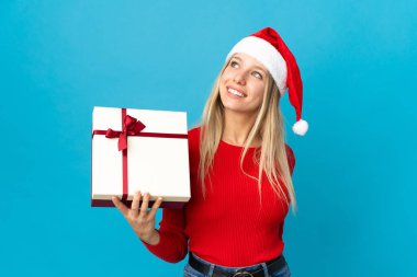 Noel şapkalı bir kadın mavi arka planda izole edilmiş bir hediyeyi tutuyor gülümserken yukarı bakıyor