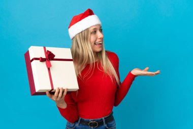 Elinde hediye tutan, Noel şapkalı bir kadın. Mavi arka planda, yüzünde sürpriz bir ifadeyle izole edilmiş.