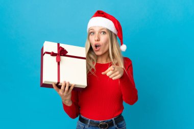Noel şapkalı kadın elinde bir hediye tutuyor mavi arka planda, şaşırmış ve ön tarafı işaret ediyor.