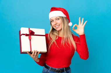 Noel şapkalı kadın mavi arka planda izole edilmiş bir hediye tutuyor. Parmaklarıyla tamam işareti gösteriyor.