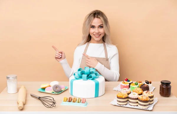 Teenager pastry chef with a big cake in a table pointing finger to the side