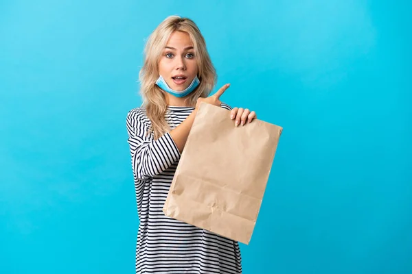 若いですロシアの女性を保持しています食料品の買い物袋青の背景に隔離驚きと指摘側 — ストック写真