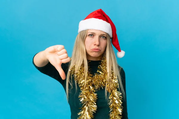 Mavi arka planda izole edilmiş noel şapkalı bir kadın olumsuz ifadeyle baş parmağını aşağı gösteriyor.