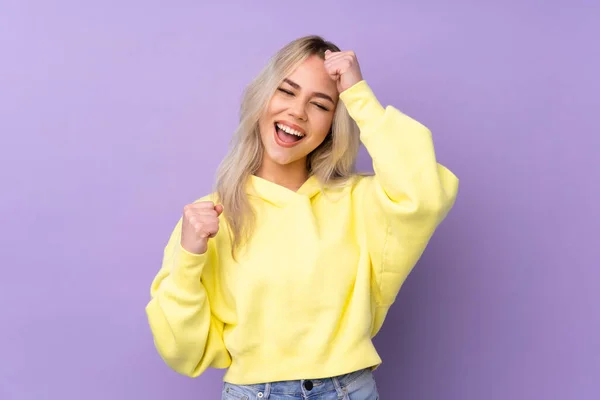 Teenager girl wearing a yellow sweatshirt over isolated purple background celebrating a victory