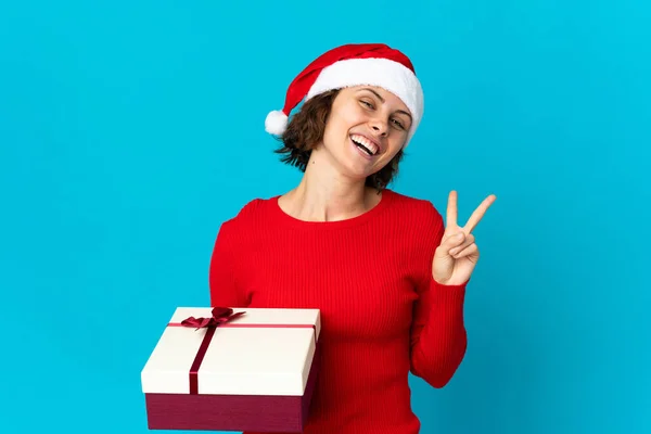 英語の女の子とともにクリスマス帽子を保持していますプレゼント孤立オン青背景笑顔とショー勝利サイン — ストック写真