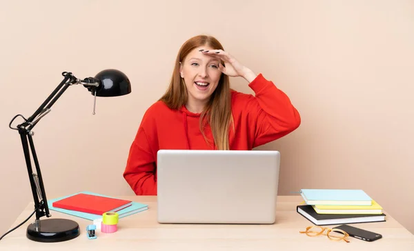 Young student woman in a workplace with a laptop looking far away with hand to look something