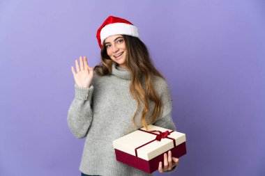 Noel şapkalı kız beyaz arka planda izole edilmiş bir hediye tutuyor mutlu bir ifadeyle selamlıyor.