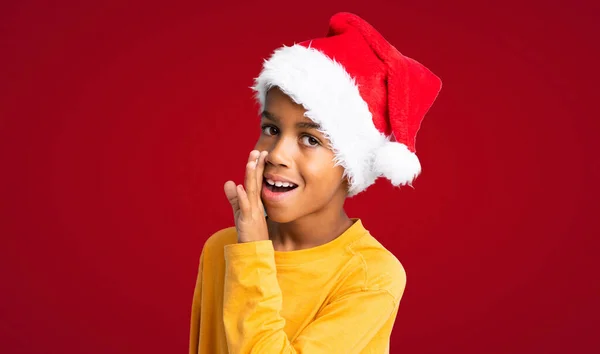 戴着圣诞礼帽的非裔美国男孩在红色背景上低声说了些什么 — 图库照片