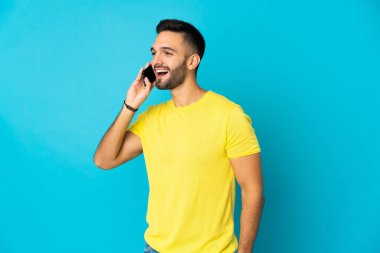 Genç beyaz adam mavi arka planda izole edilmiş cep telefonuyla sohbet ediyor.