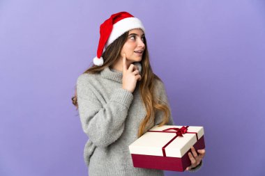 Noel şapkalı kız elinde hediyeyle beyaz arka planda izole edilmiş bir şekilde yukarı bakarken bir fikir düşünüyor.