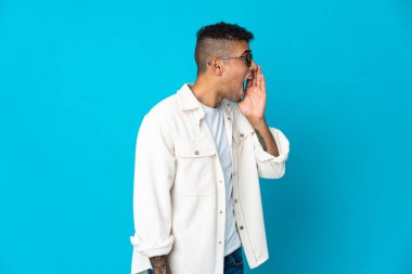 Mavi arka planda izole edilmiş genç Brezilyalı adam ağzı sonuna kadar açık bağırıyor.