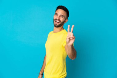 Mavi arka planda izole edilmiş genç beyaz adam gülümsüyor ve zafer işareti gösteriyor.