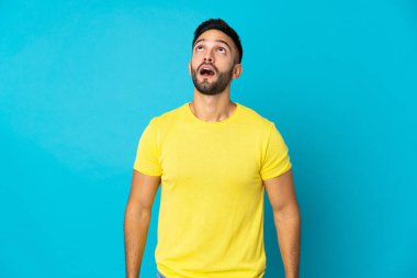 Kafkasyalı genç adam mavi arka planda izole edilmiş yukarı bakıyor ve şaşırmış bir ifadeyle