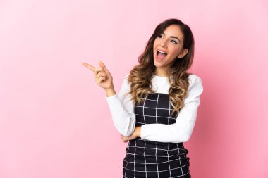 Pembe arka planda izole edilmiş genç beyaz bir kadın parmak göstererek bir ürün sunuyor.
