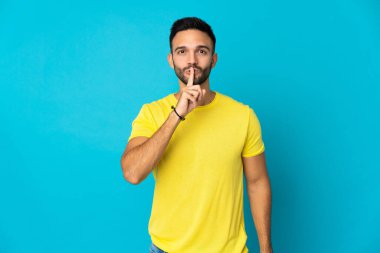 Mavi arka planda izole edilmiş genç beyaz adam sessiz bir jest işareti gösteriyor. Parmağını ağzına sokuyor.
