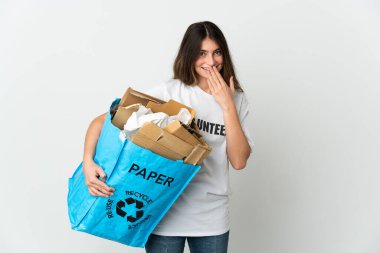 Elinde kağıt dolu bir geri dönüşüm çantası tutan genç bir kadın beyaz arka planda izole edilmiş mutlu ve gülümseyen bir ağızla elini kapatıyor.