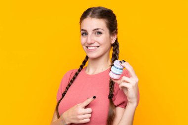 Soyutlanmış sarı arka planda genç bir kadın elinde renkli Fransız kurabiyeleriyle onu işaret ediyor.