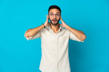 Mavi arka planda izole edilmiş genç beyaz adam hayal kırıklığına uğramış ve kulaklarını kapatmış.