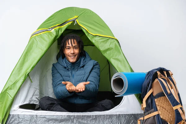 Young african american man inside a camping green tent holding copyspace imaginary on the palm to insert an ad