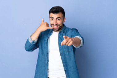 İzole edilmiş mavi arka plan üzerinde beyaz bir adam telefon hareketi yapıyor ve ön tarafı işaret ediyor.