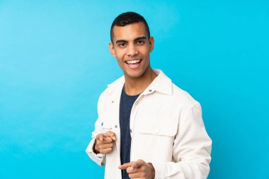 Genç Afro-Amerikalı, izole edilmiş mavi arka planda ön tarafı işaret ediyor ve gülümsüyor.