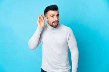 İzole edilmiş mavi arka planda beyaz bir adam. Kulağına elini sürerek bir şeyler dinliyor.