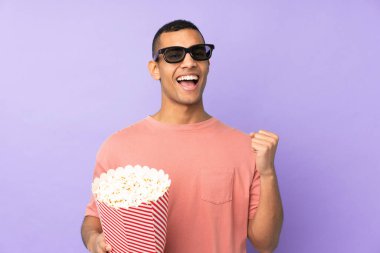 Üç boyutlu gözlüklü ve elinde koca bir kova patlamış mısırla izole edilmiş mavi arka planı olan genç bir Afro-Amerikalı.