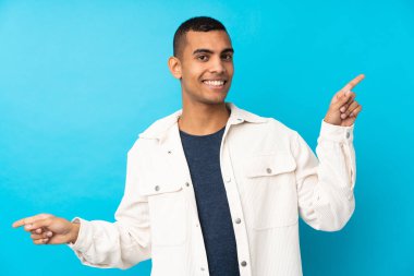 İzole edilmiş mavi arka planda genç bir Afro-Amerikan erkeği parmak uçlarını işaret ediyor ve mutlu