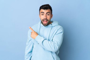 İzole edilmiş mavi arka planı olan beyaz bir adam şaşırmış ve işaret ediyor.