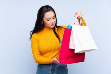 İzole edilmiş mavi arka planı olan genç Kolombiyalı kız elinde alışveriş çantaları tutuyor.