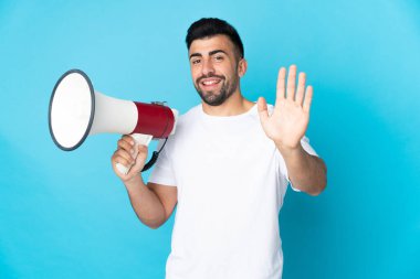 İzole edilmiş mavi arka plan üzerinde beyaz bir adam elinde bir megafon ve mutlu bir ifadeyle selam veriyor.