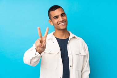 Genç Afrikalı Amerikalı adam mavi arka planda gülümsüyor ve zafer işareti gösteriyor.