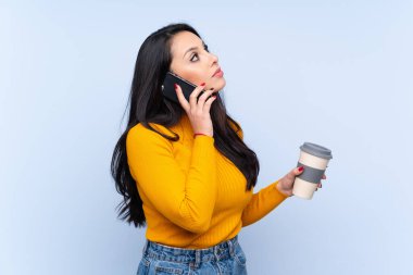 İzole edilmiş mavi arka planda genç bir Kolombiyalı kız elinde kahve ve bir cep telefonu tutuyor.