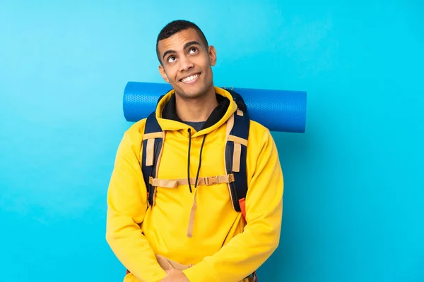 孤立した青の背景に大きなバックパックを持つ若い登山家の男笑いと検索アップ — ストック写真