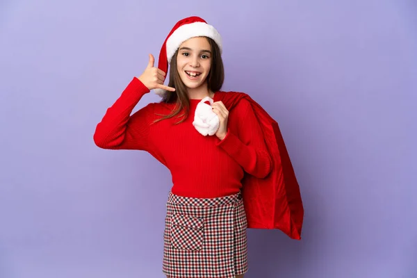 Little girl with hat and Christmas sack isolated on purple background making phone gesture. Call me back sign