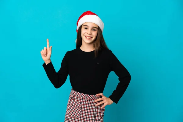 戴着圣诞礼帽的小女孩被蓝色的背景隔开 她举起一个手指 表示她是最好的象征 — 图库照片
