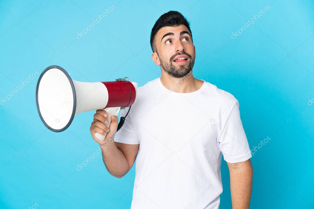 Caucasian man over isolated blue background holding a megaphone and looking up while smiling