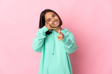 Pembe arka planda izole edilmiş genç Latin bir kadın telefon hareketi yapıyor ve ön tarafı işaret ediyor.