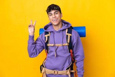 Sarı arka plan üzerinde büyük bir sırt çantası olan genç dağcı gülümsüyor ve zafer işareti gösteriyor.