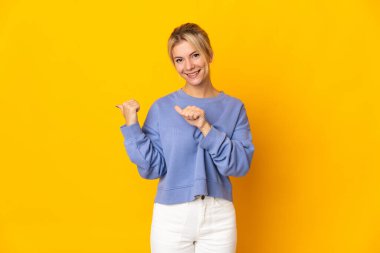Sarı arka planda izole edilmiş genç bir Rus kadın bir ürün sunmak için kenarı işaret ediyor.