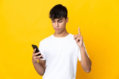 Young man over isolated yellow background using mobile phone and lifting finger