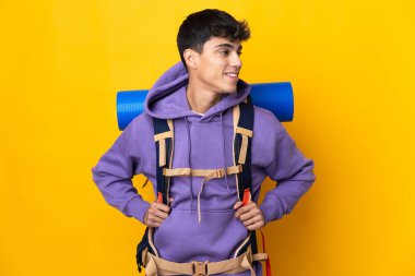Young mountaineer man with a big backpack over isolated yellow background looking side