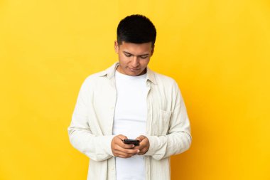 Genç Ekvadorlu, sarı arka planda izole edilmiş cep telefonuyla mesaj gönderiyor.