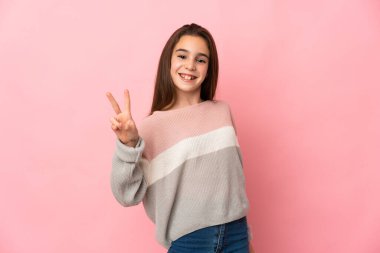 Pembe arka planda izole edilmiş küçük kız gülümsüyor ve zafer işareti gösteriyor