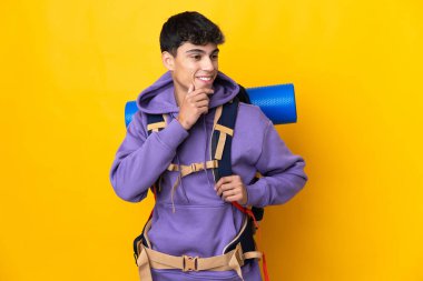 Young mountaineer man with a big backpack over isolated yellow background looking to the side and smiling