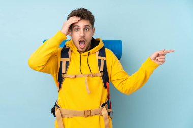 Young mountaineer man with a big backpack isolated on blue  background surprised and pointing finger to the side